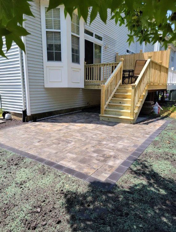 A newly built wooden deck with stairs leading to a paver patio, next to a light gray house.