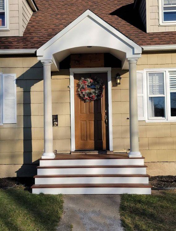 Beige house with wooden door, steps, and columns supporting a white awning; wreath on the door.