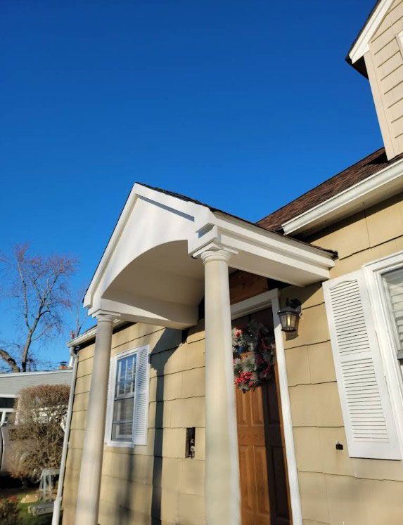 Beige house exterior with a porch, two columns, brown door, wreath, white shutters, and clear blue sky.