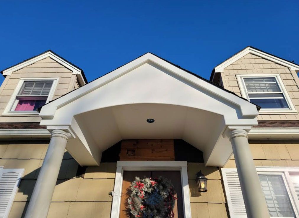 Two-story house with beige siding, white trim, and blue sky. Porch has columns and a wreath on the door.