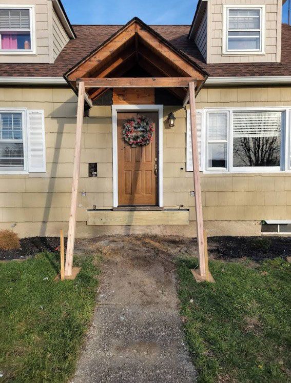 A newly constructed wooden porch frame outside a tan house with a brown door and a welcome wreath.