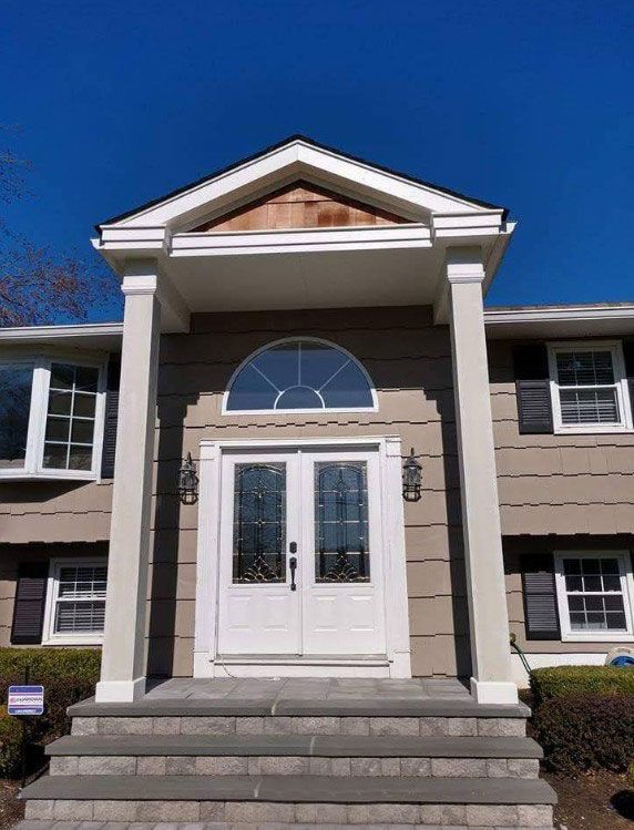 White double doors under a portico with gray pillars and steps; house with brown siding, blue sky.
