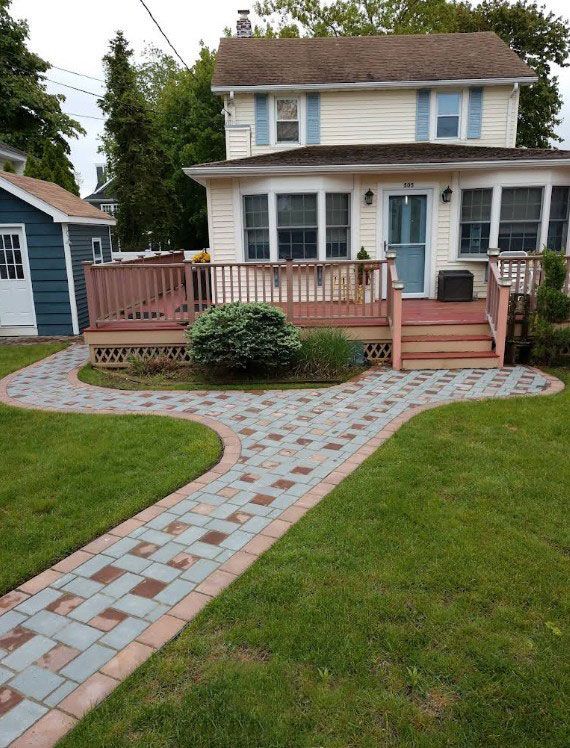 Brick walkway leads to a two-story house with a deck, a green lawn, and a blue shed.