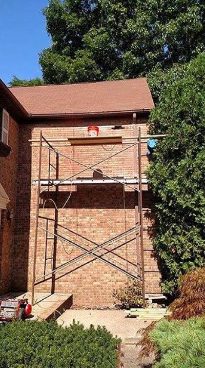 Brick wall with scaffolding, a person working, and a red bucket, next to greenery and roof.