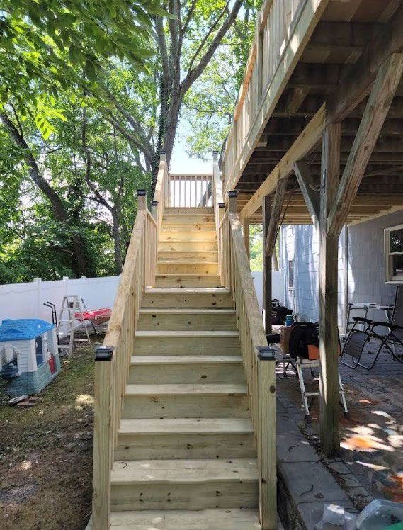 Wooden staircase leading up to a deck, viewed from the ground. Surrounded by trees and a backyard.