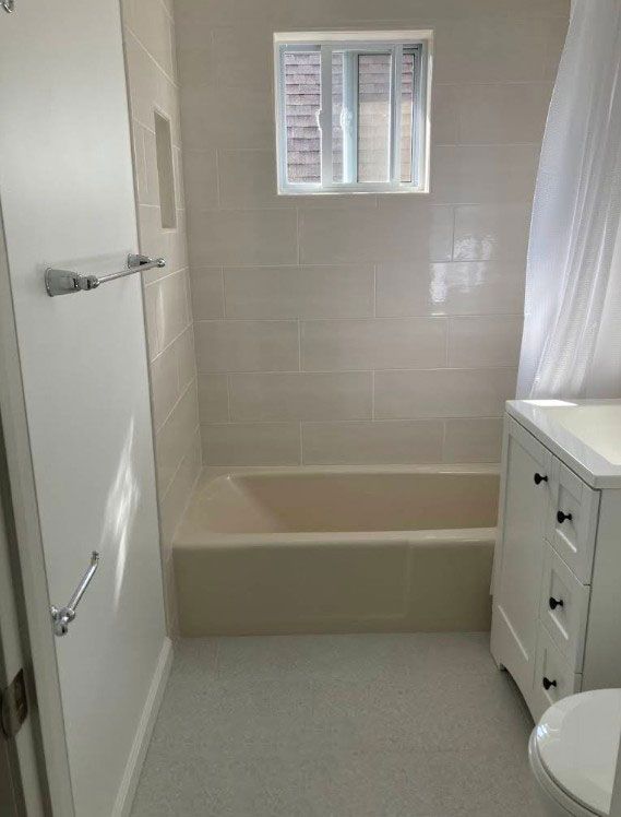 Small bathroom with beige tub, white tiles, vanity, and light gray floor. Small window above the tub.