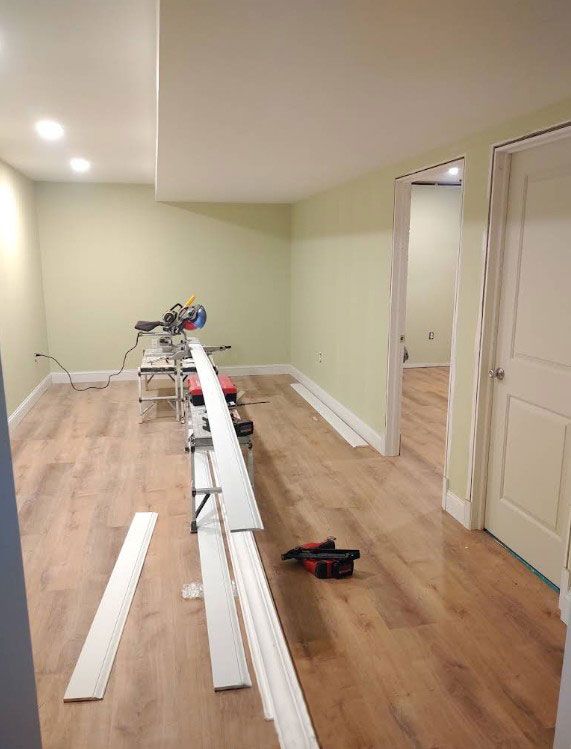 Room renovation in progress; baseboards laid out, miter saw, light green walls, wood-look floor.