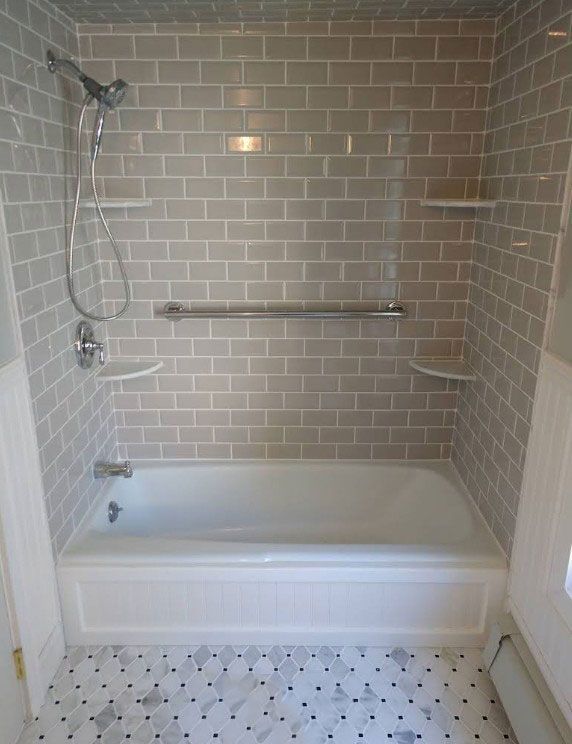 A white tub/shower with gray tile walls, white shelves, and a silver handrail.