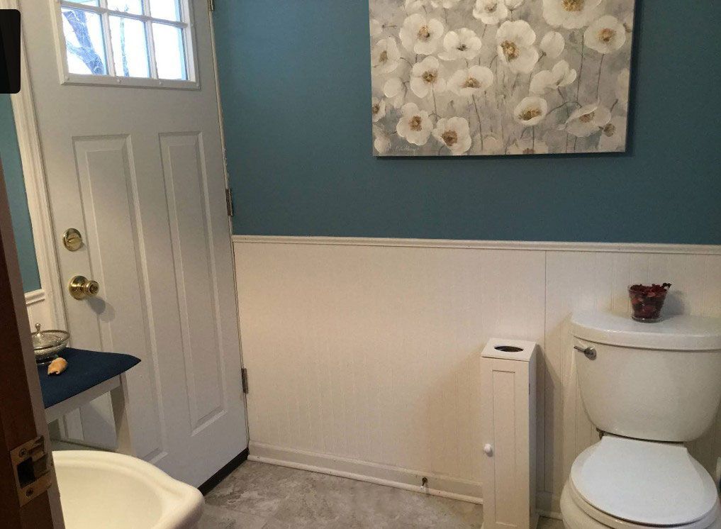 Bathroom with white wainscoting, blue wall, floral art, and toilet. A white door with a window is visible.