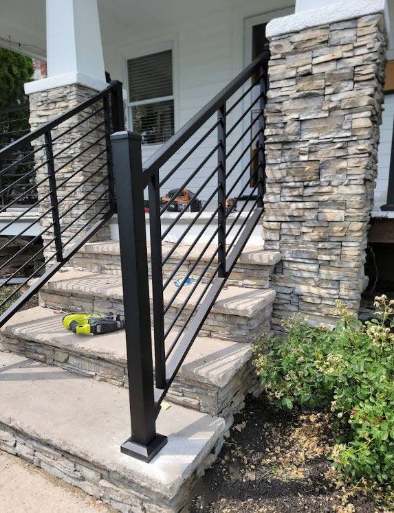 Black metal handrail on stone steps of a house with stone-covered columns and landscaping.