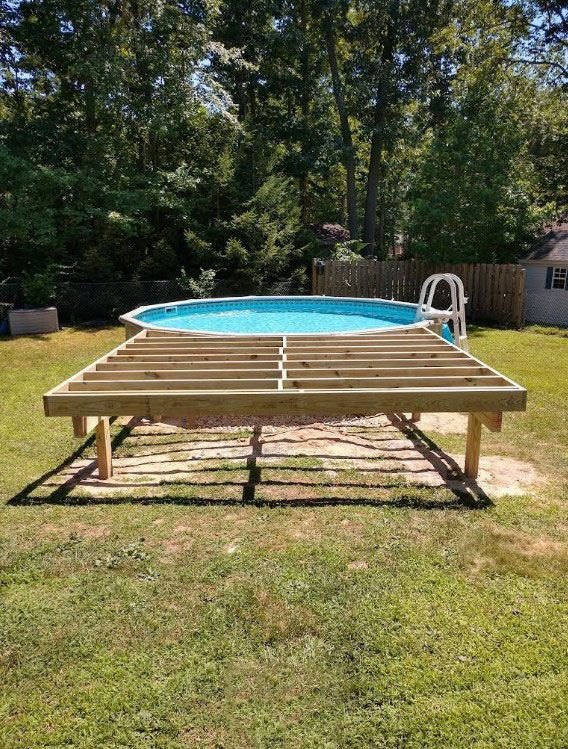 Above-ground pool on wooden deck in a backyard, surrounded by grass and trees on a sunny day.