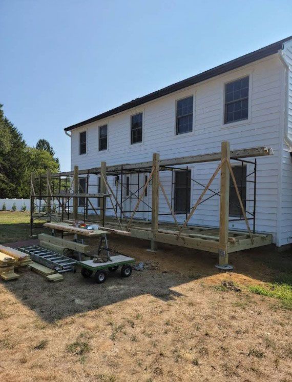 Deck construction in progress next to a two-story white house. Scaffolding and lumber are visible.