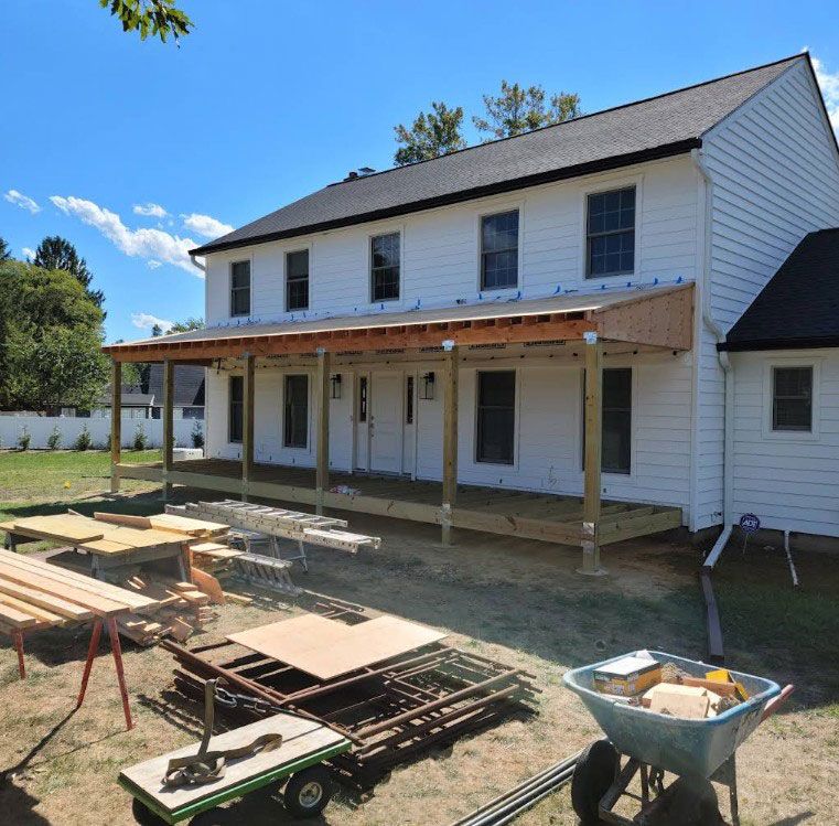 House with a new wooden porch under construction; white siding, green grass, blue sky, construction materials.