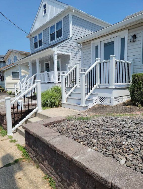 Two-story gray house with white trim and steps; brown retaining wall in front; sunny outdoor setting.