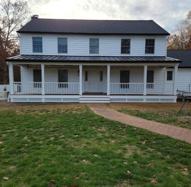 White two-story house with a wraparound porch and dark roof, set in a grassy yard.