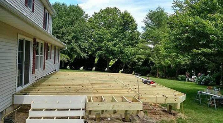 Newly built wooden deck attached to a house, with stairs and tools visible. Green trees in background.