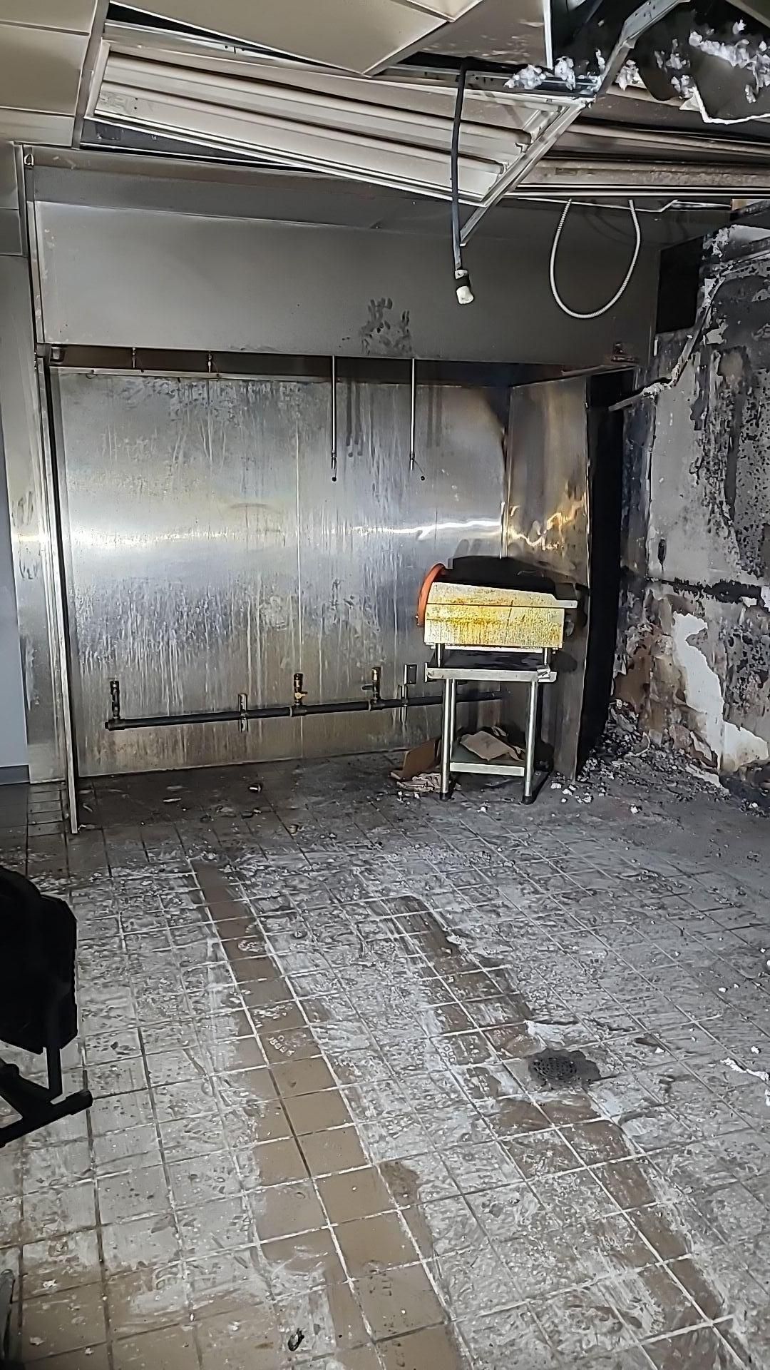 Fire-damaged interior of a room, walls blackened. A stainless steel structure with a yellow cart visible. Debris covers the floor.