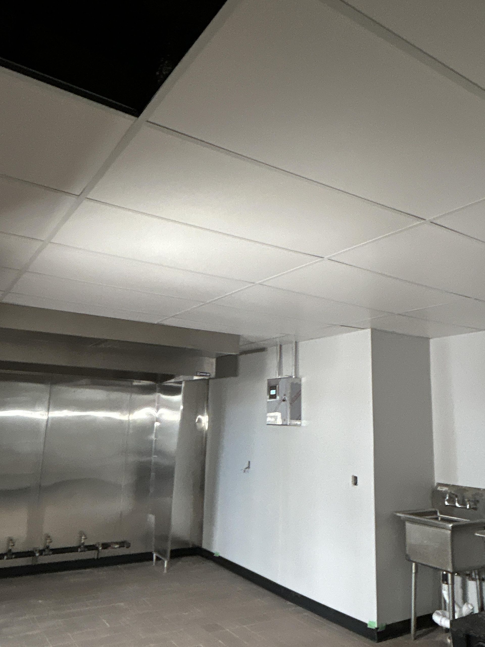 Commercial kitchen interior with stainless steel equipment, white walls, and a tiled ceiling.