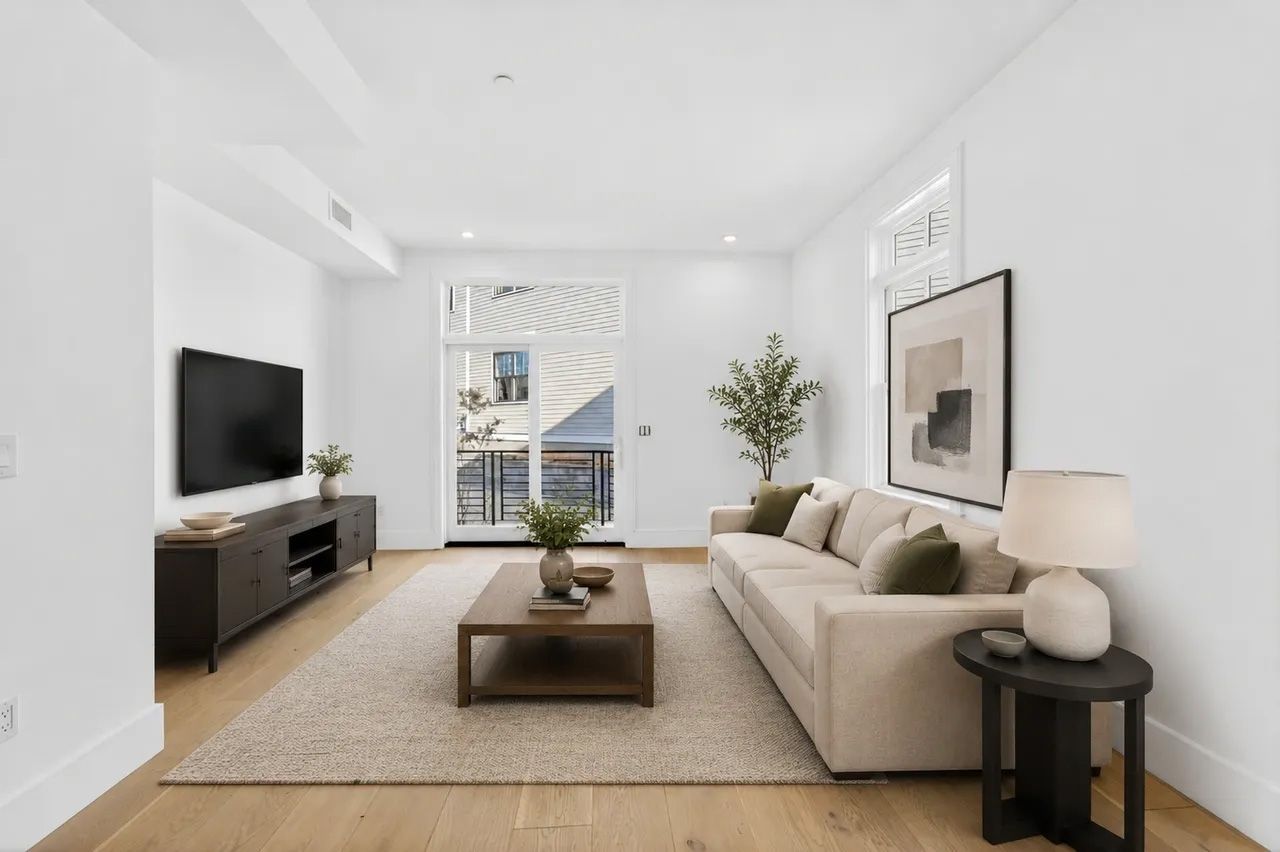 Bright modern living room with beige sofa, TV, coffee table, and large window overlooking a street.