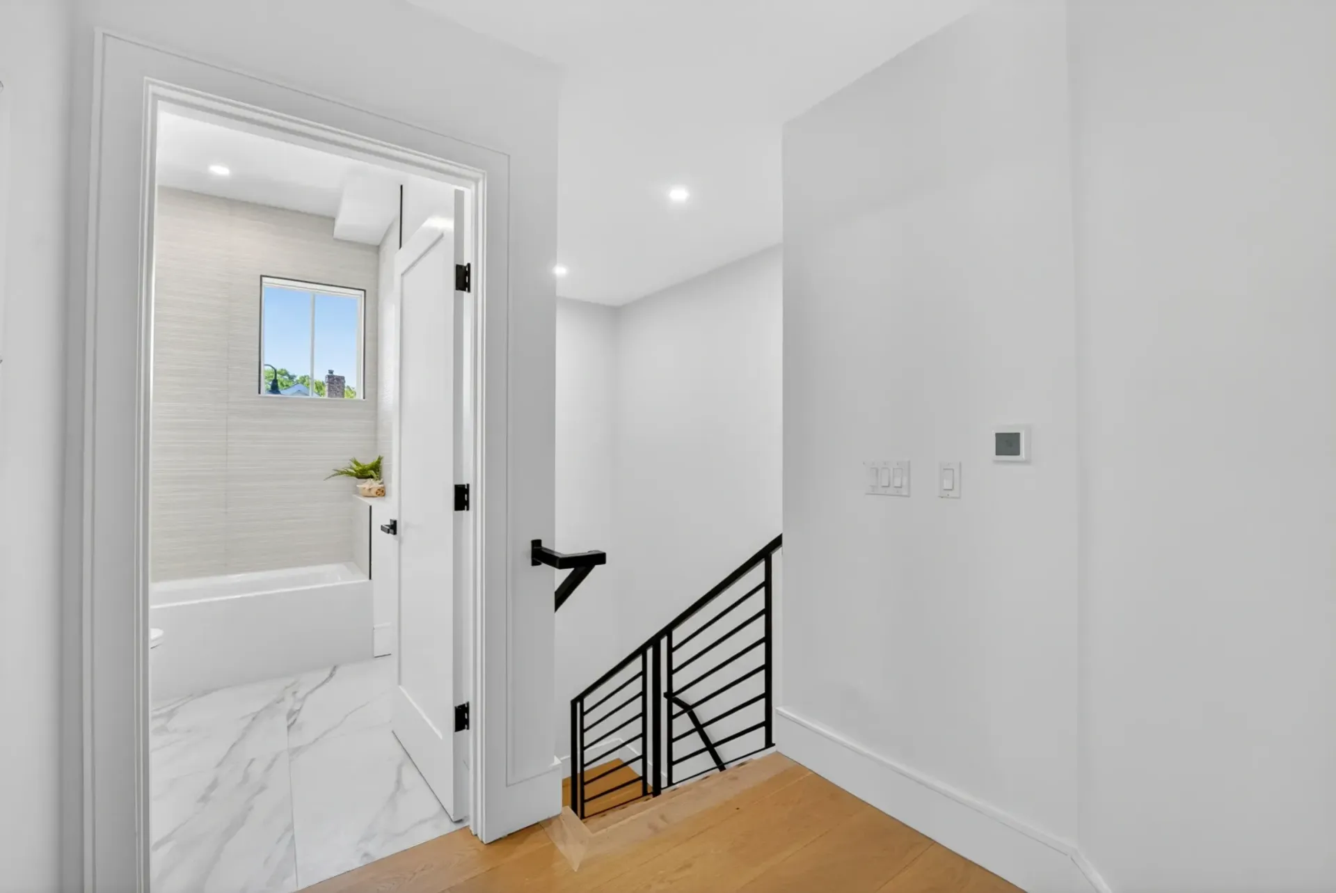 Bright white hallway with open bathroom door, staircase railing, and light wood floor, viewed from upstairs.