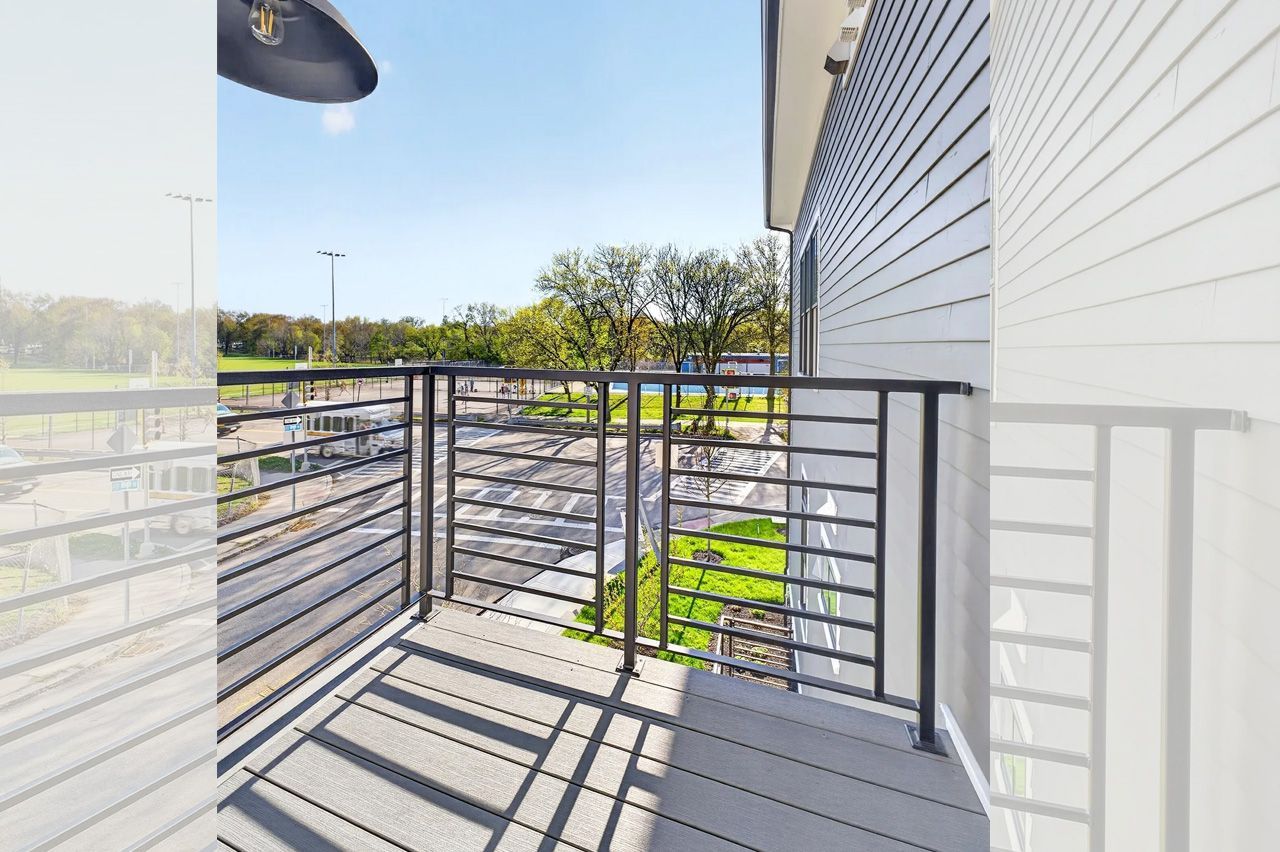 Balcony with metal railing beside a white building, overlooking a green courtyard on a sunny day