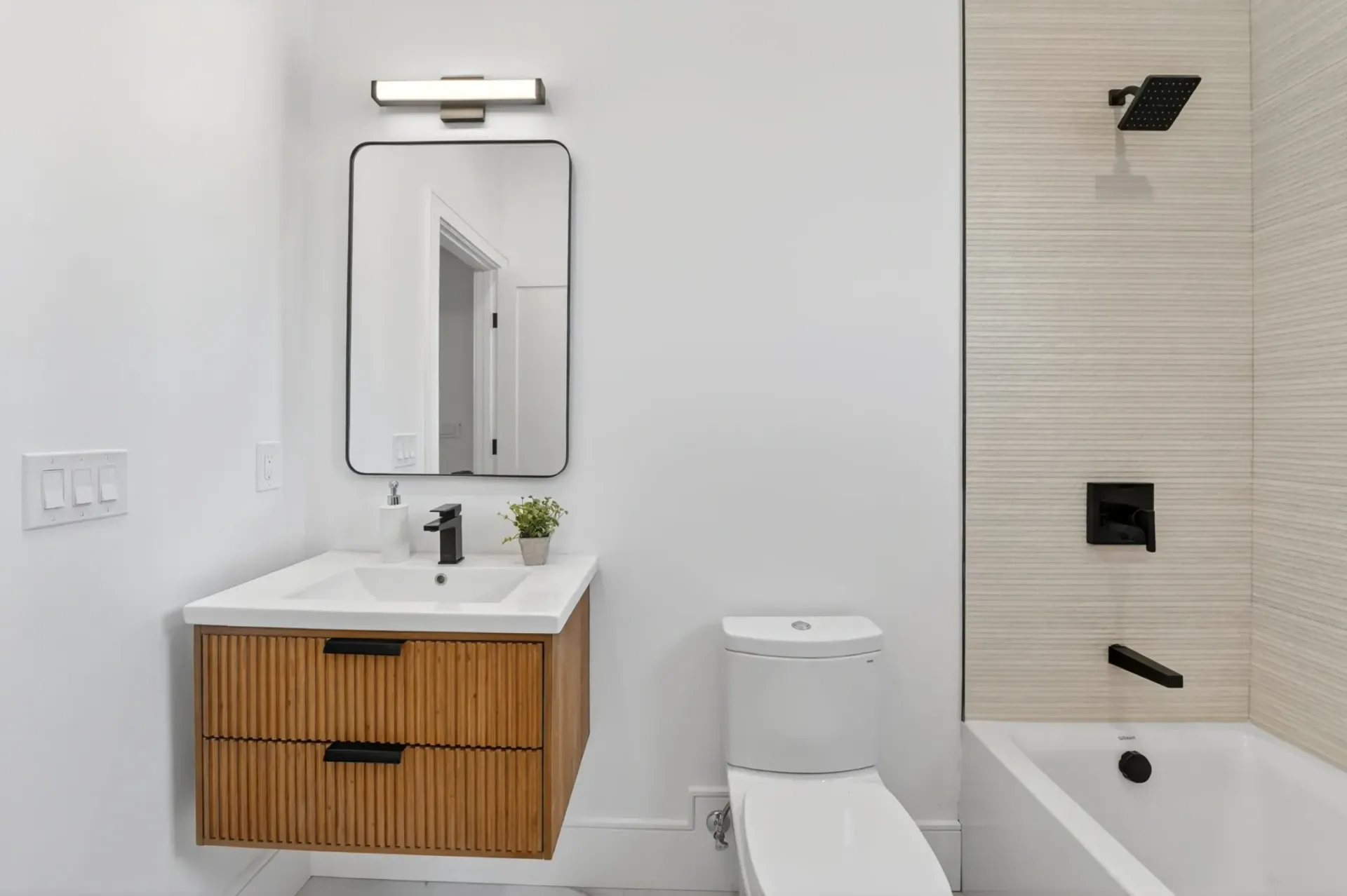 Modern bathroom with wood vanity, mirror, toilet, and beige tiled bathtub shower.
