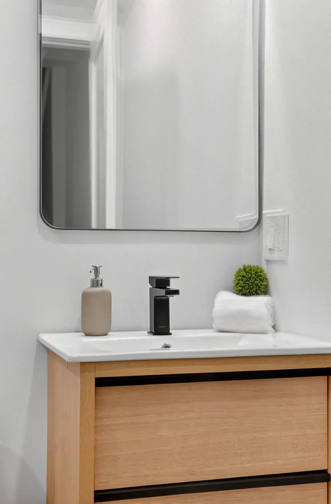 Modern bathroom vanity with wood cabinet, white countertop, black faucet, mirror, soap dispenser, and small plant.