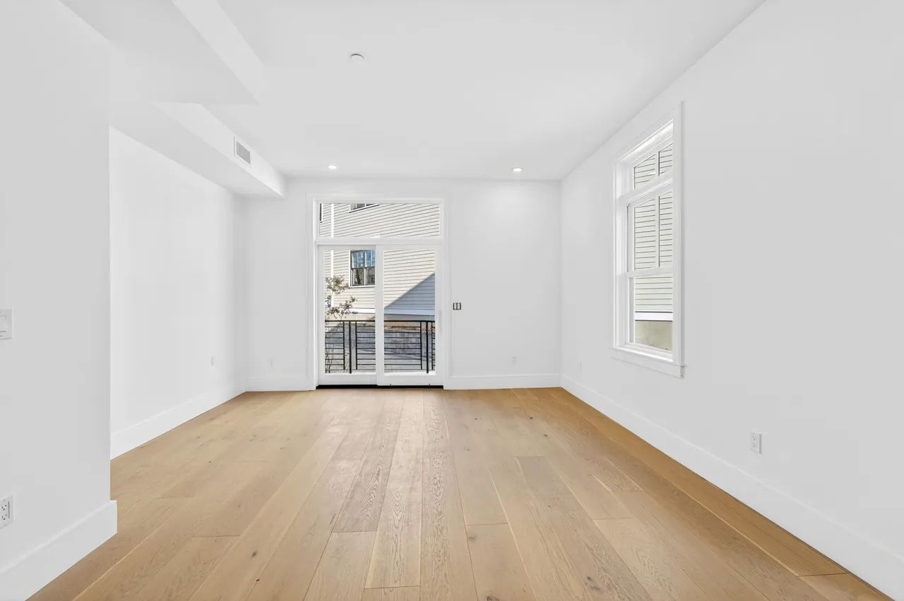 Bright empty room with light wood floors, white walls, and a glass door to a balcony.