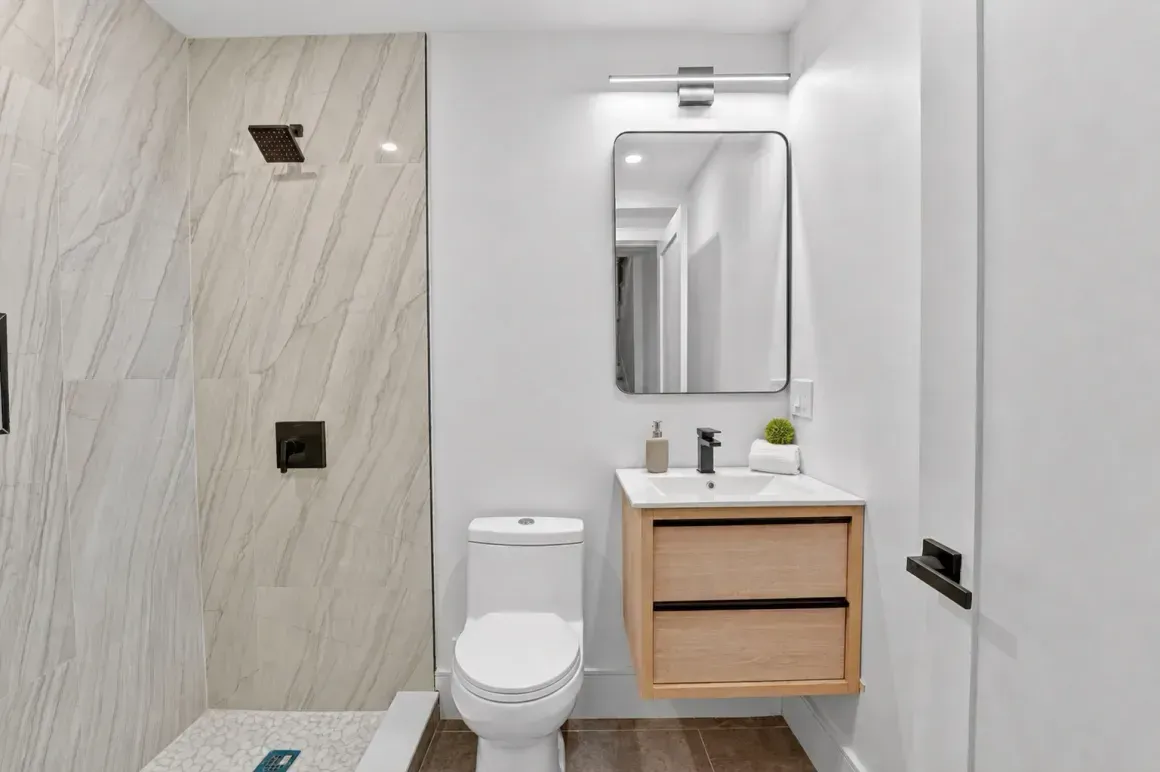 Modern bathroom with toilet, wood vanity, mirror, and tiled shower wall