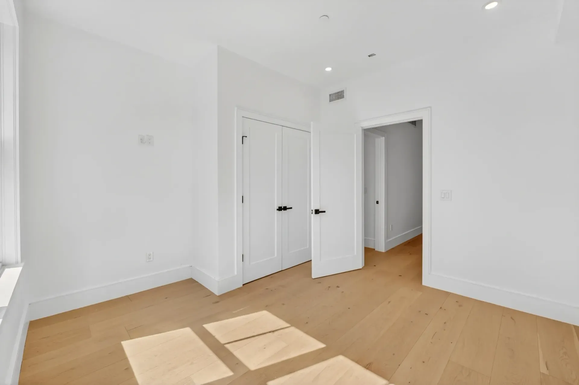 Bright empty white room with wood floor and double closet doors, lit by sunlight from a window.