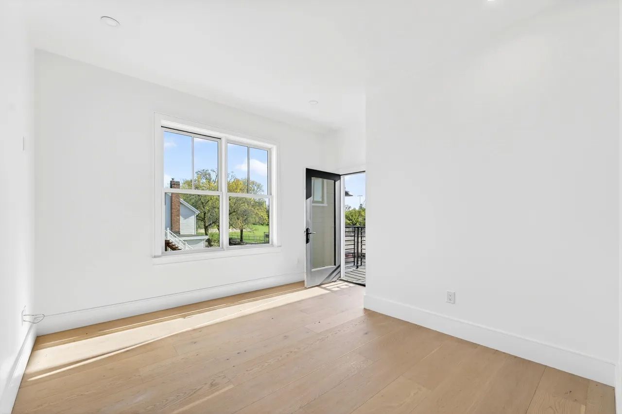A bright empty room with white walls, light wood floor, and a large window overlooking greenery