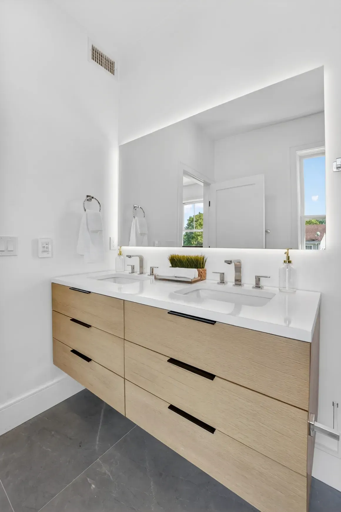 Modern bathroom vanity with light wood drawers, white countertop, large mirror, and gray tile floor
