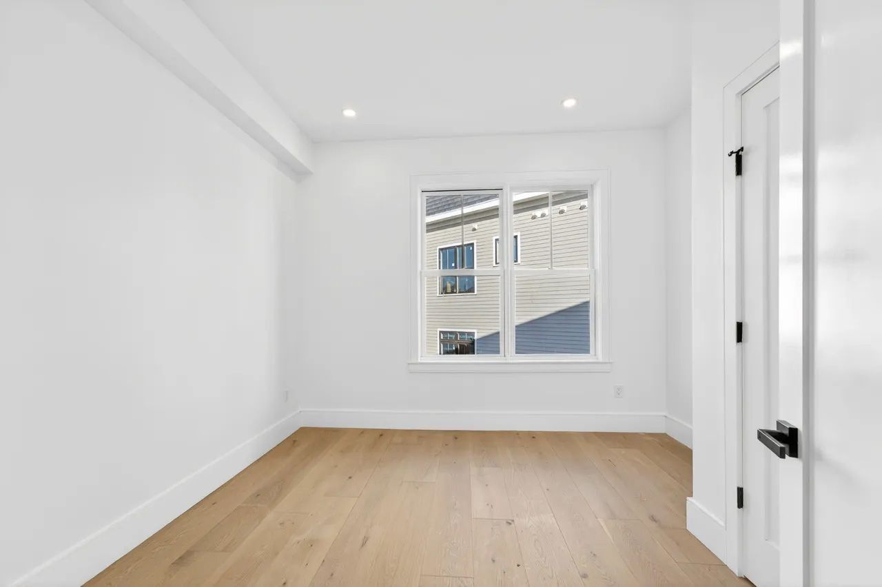 Empty white room with light wood floors, a window, recessed lights, and a door on the right