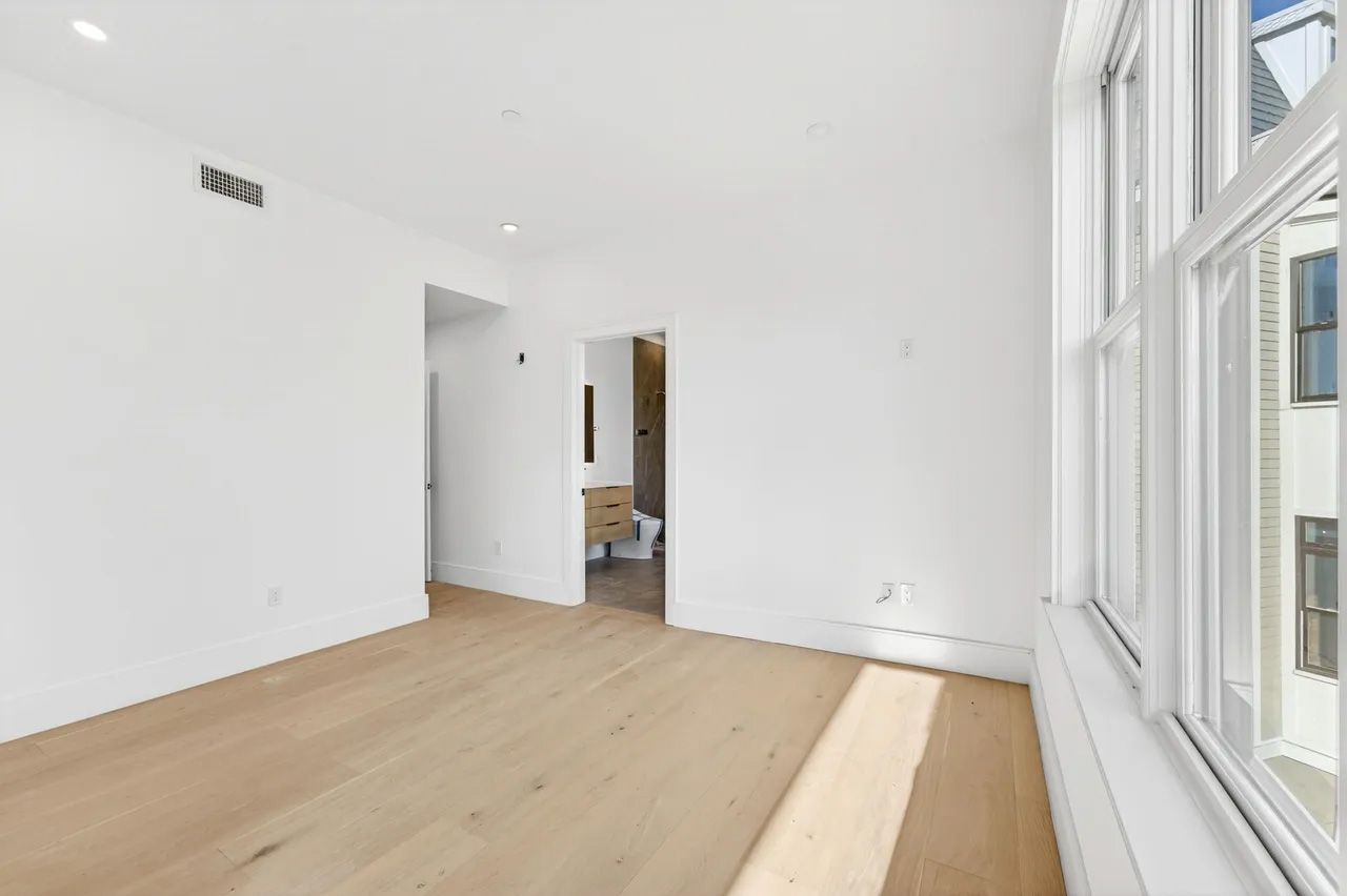 Bright empty room with white walls, light wood floors, and tall windows.