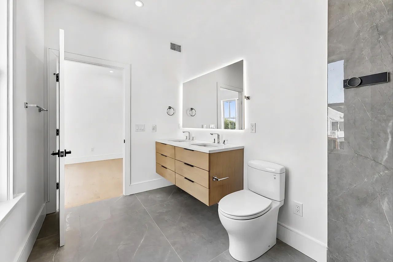 Modern white bathroom with gray tile, wood vanity, mirror, toilet, and shower.