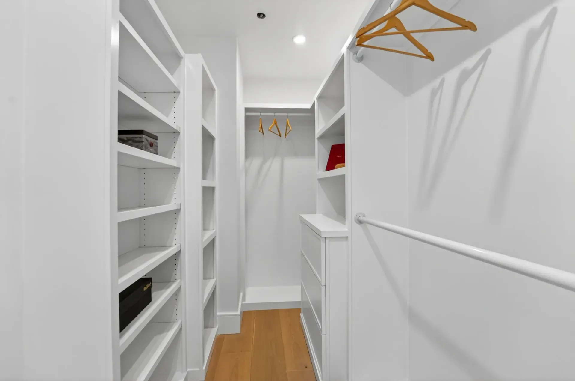Bright walk-in closet with white built-in shelves, hanging rods, and wooden floorboards
