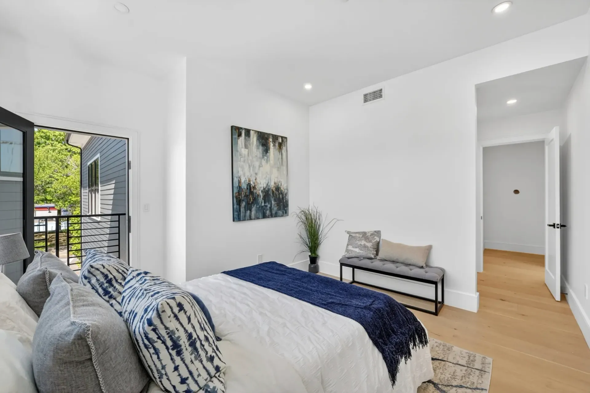 Bright modern bedroom with bed, bench, wall art, and open balcony doors