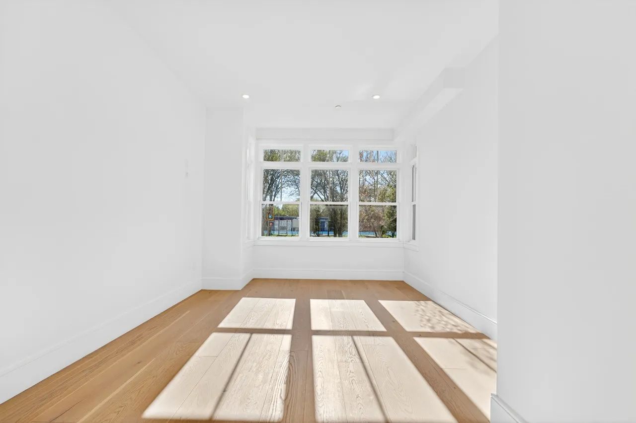 A bright empty room with white walls, wood floor, and large windows casting sunlight and shadows