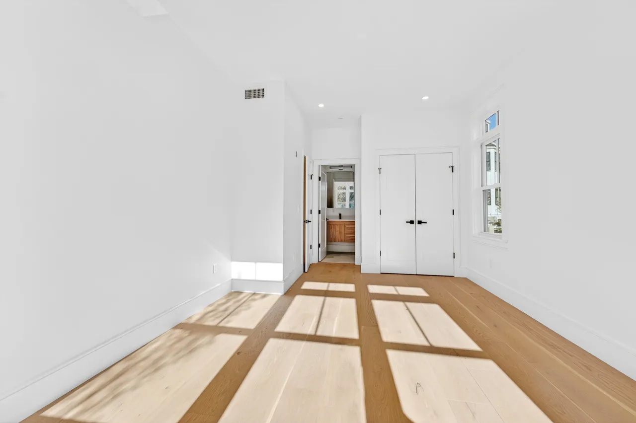 A bright empty room with white walls, wood floors, and sunlight casting window shadows.