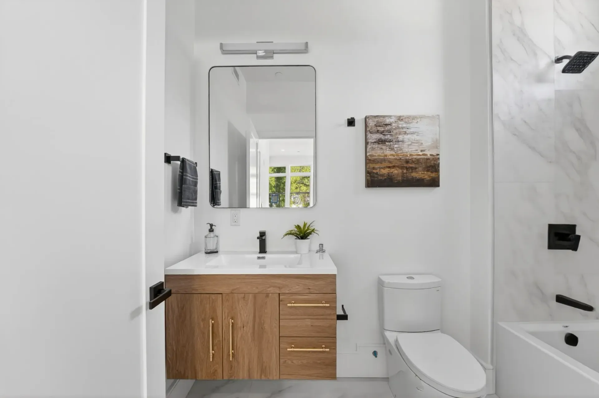 Bright modern bathroom with white walls, wood vanity, mirror, toilet, and tub shower.