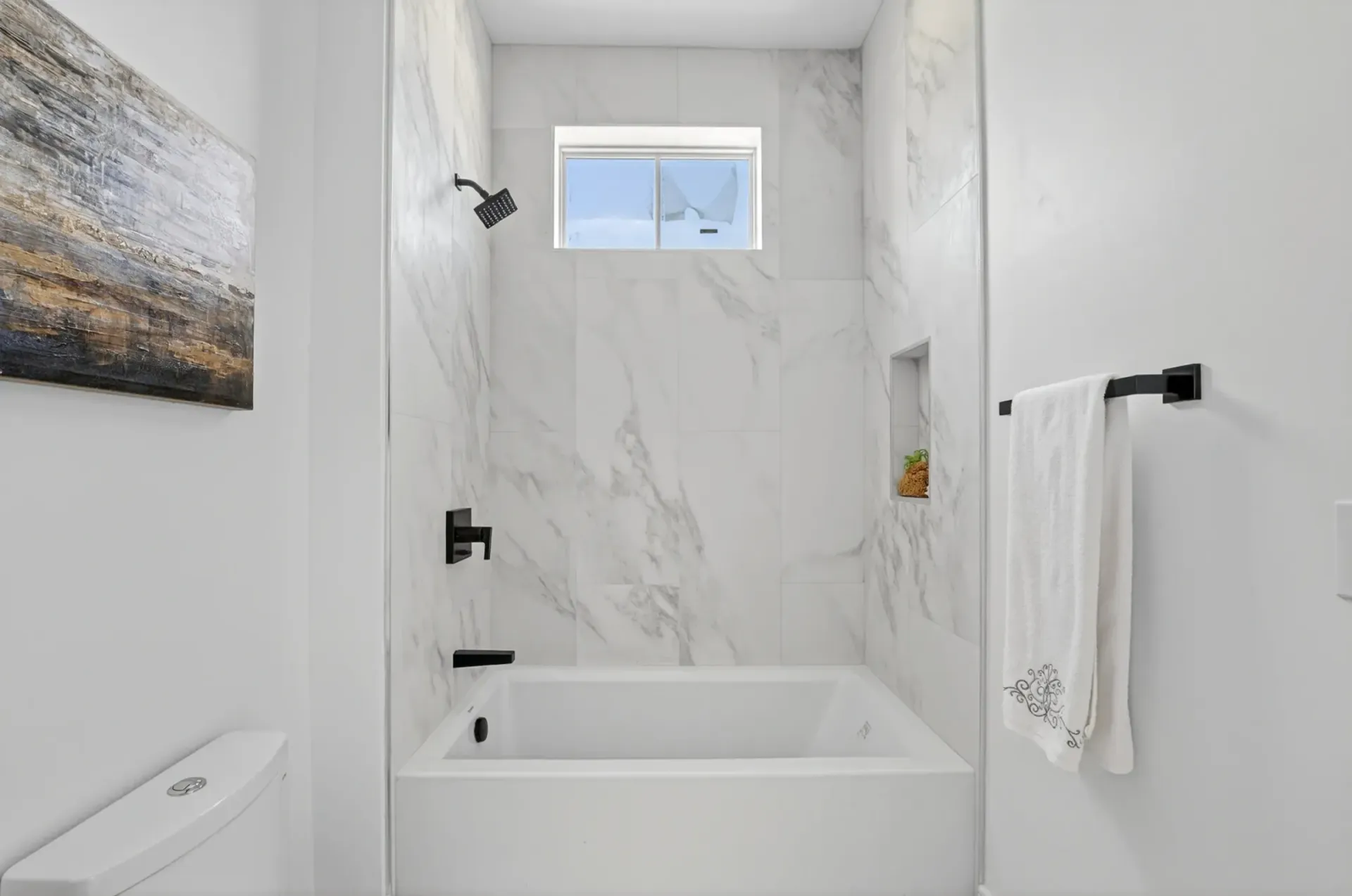 Bright white bathroom with a bathtub, marble walls, window, towel rack, and wall art.