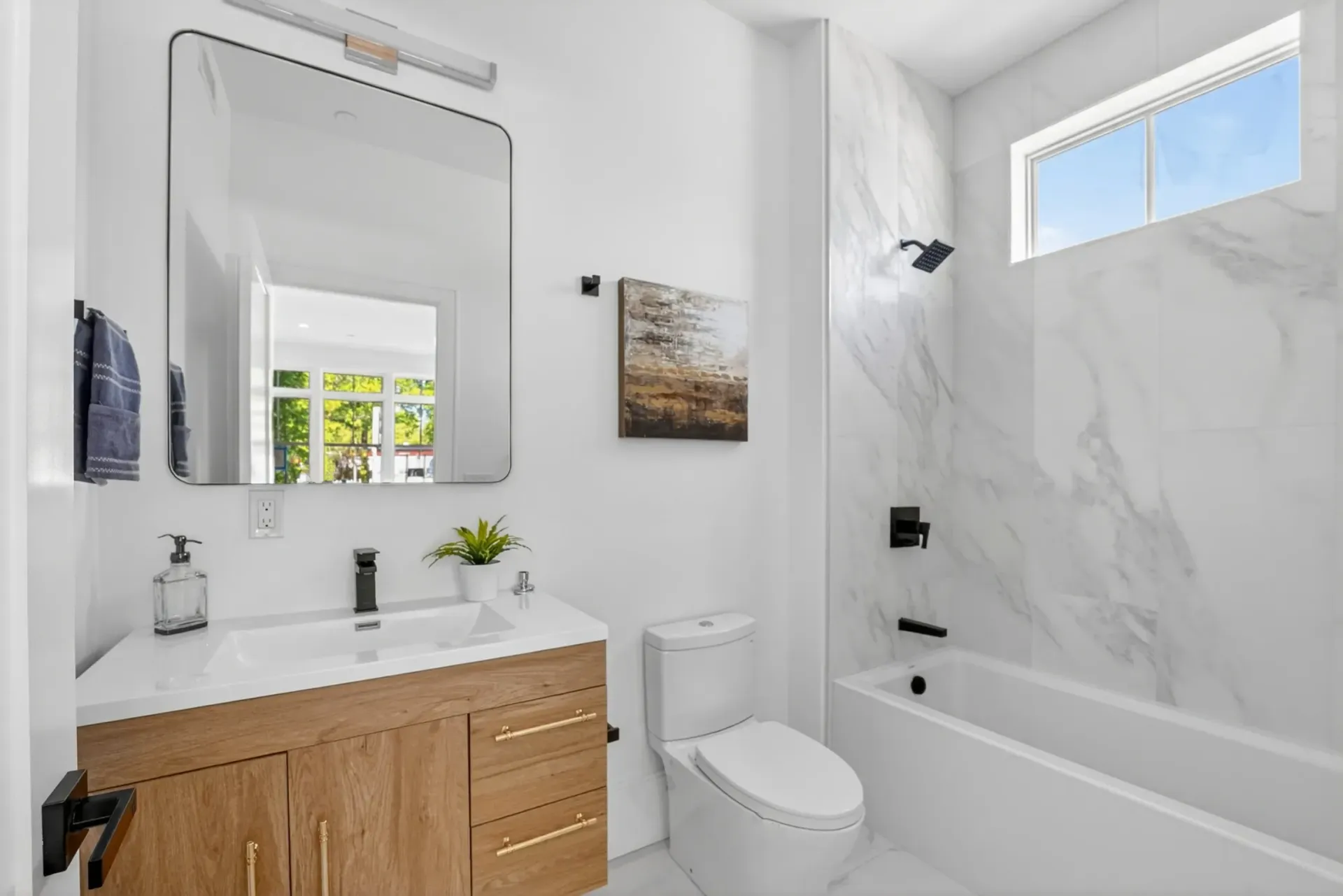 Bright modern bathroom with wood vanity, mirror, toilet, and tiled bathtub/shower.