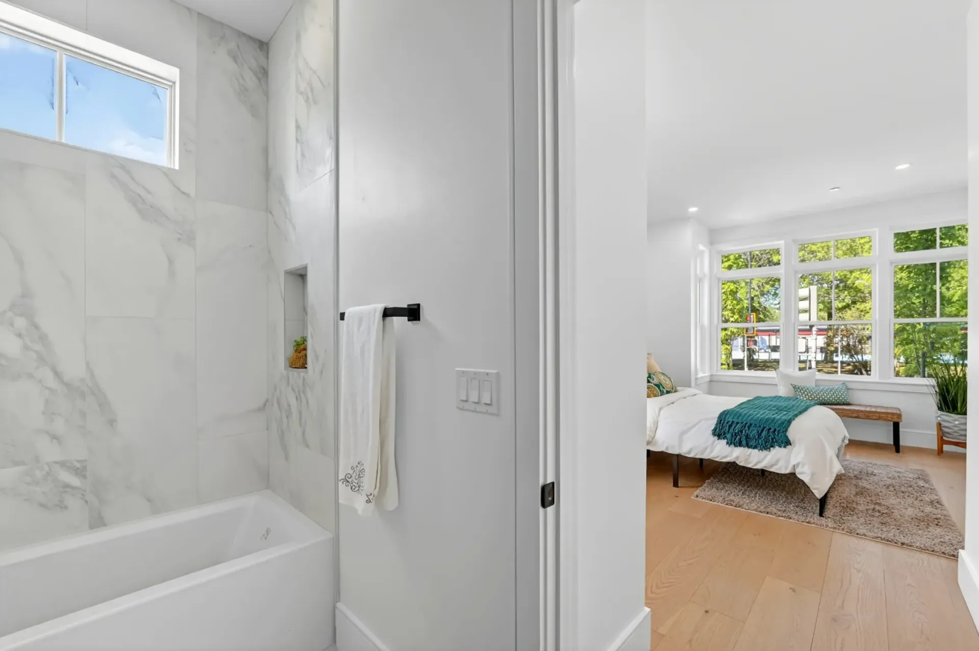 Bright bathroom with marble shower, open door to sunlit bedroom with large windows and bed