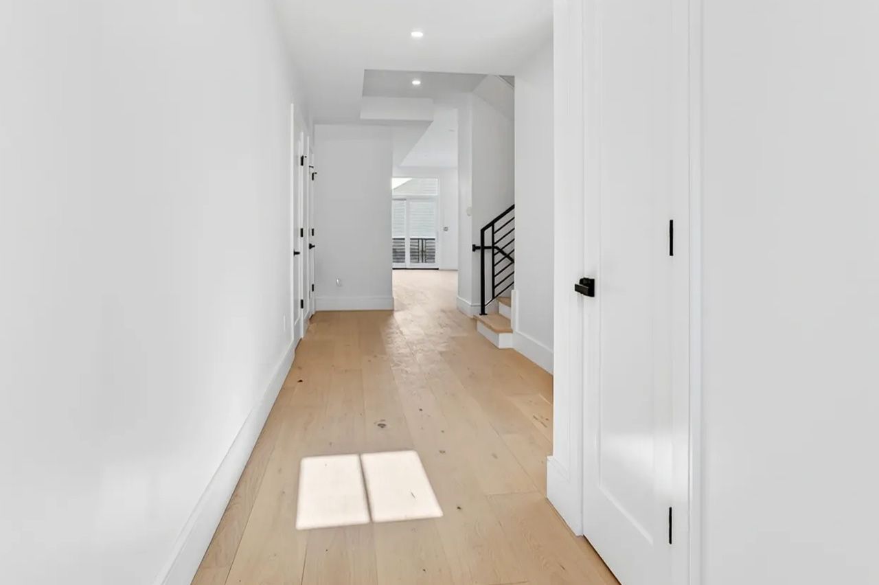 Bright hallway with white walls, light wood floors, and a staircase in the distance