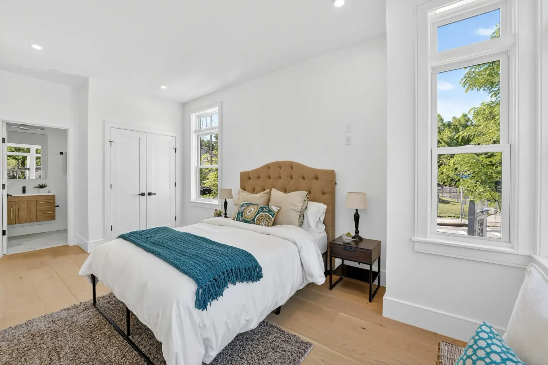 Bright bedroom with tufted bed, white walls, teal throw, and tall windows letting in daylight.