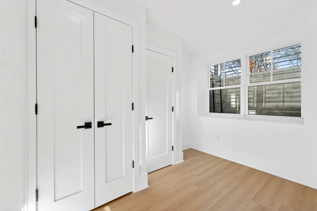 A bright empty room with white closets, white walls, a wooden floor, and two windows.