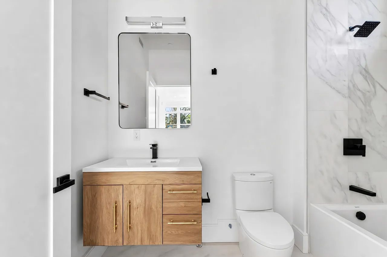 Modern white bathroom with wood vanity, mirror, sink, toilet, and marble tub surround
