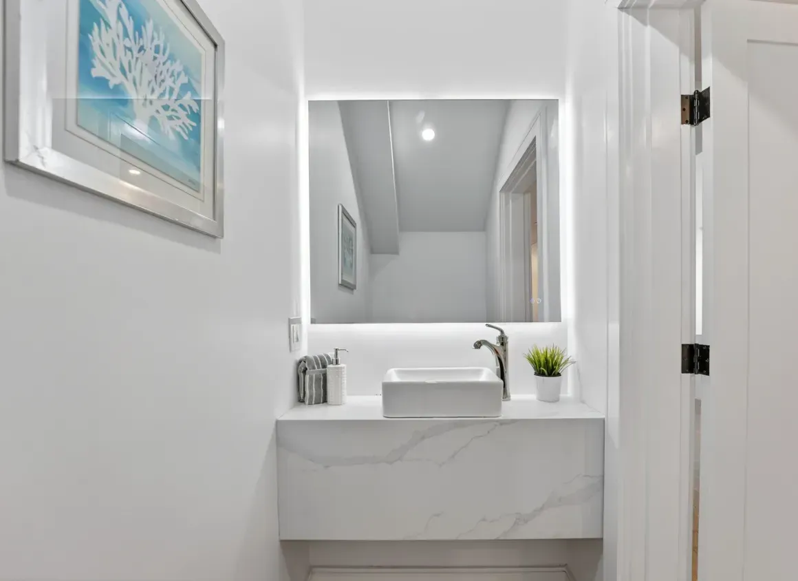 Bright white bathroom vanity with marble sink, mirror, and small plant in a narrow hallway.
