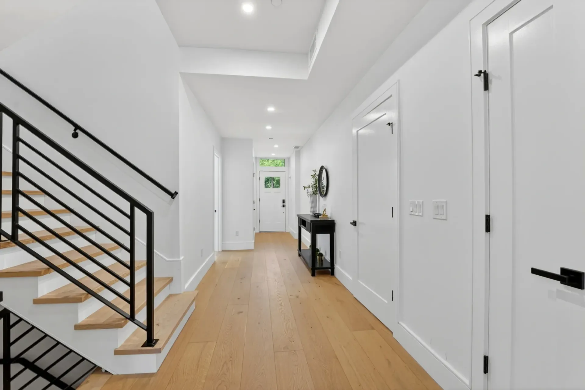 Bright white hallway with wood floors, staircase on left, and closed doors along the right wall.