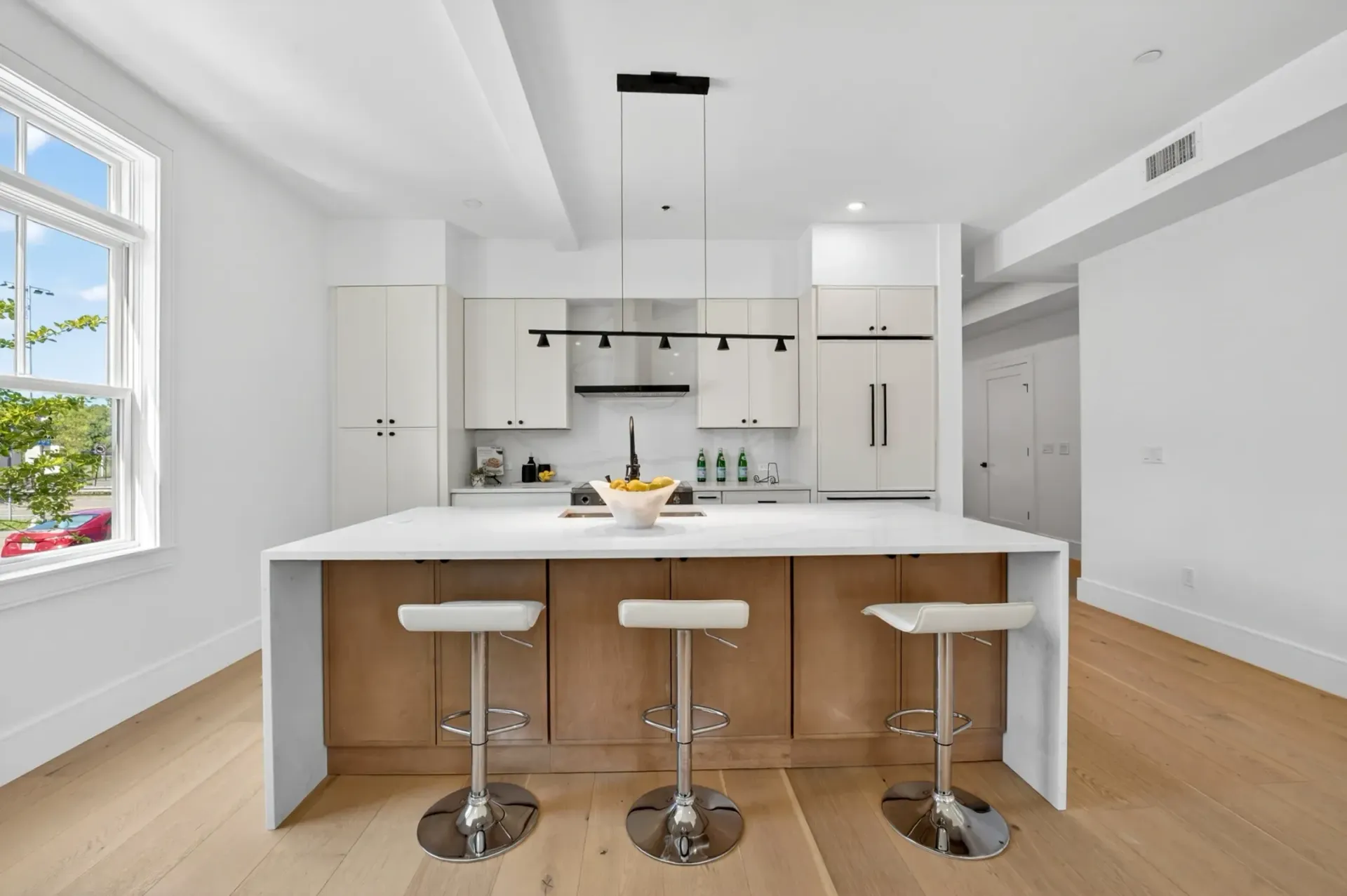 Bright modern white kitchen with island, bar stools, and wood floors, lit by large windows